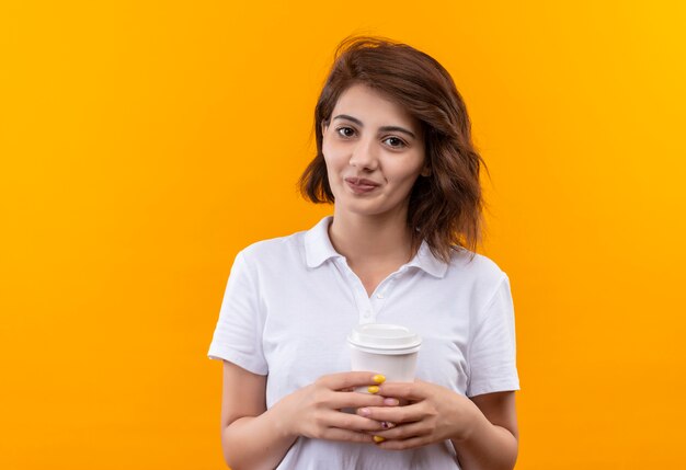 Niña con pelo corto vistiendo polo blanco sosteniendo la taza de café mirando a la cámara sonriendo con cara feliz