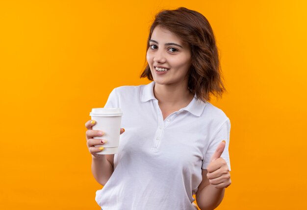 Niña con pelo corto vistiendo polo blanco sosteniendo la taza de café sonriendo alegremente mostrando los pulgares para arriba