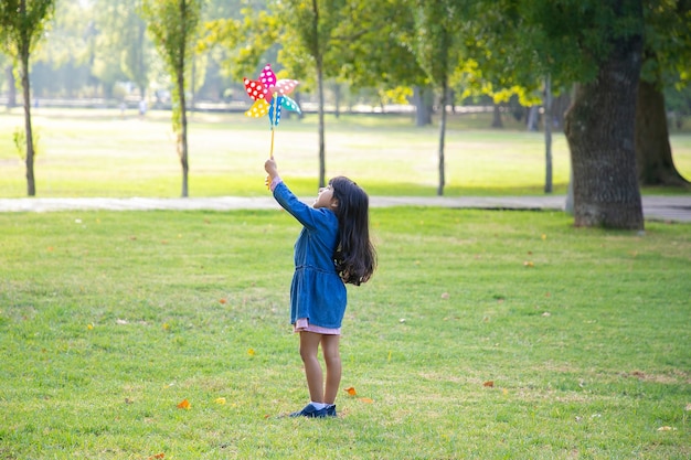 Niña de pelo negro de pie sobre el césped en el parque, sosteniendo y levantando el molinillo, mirando el juguete. Plano amplio y de longitud completa. Concepto de actividad al aire libre para niños