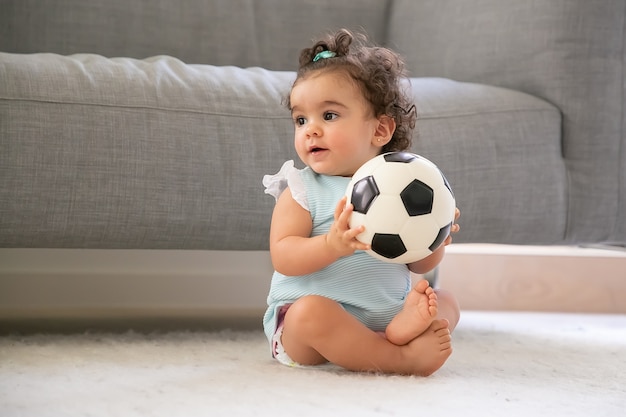 Niña de pelo negro positiva en ropa azul pálido sentada en el suelo en casa, mirando a otro lado, jugando a la pelota de fútbol. Copie el espacio. Niño en casa y concepto de infancia.