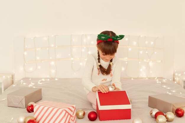 Niña de pelo oscuro con suéter blanco y cuernos de ciervo verde de fiesta abriendo caja de regalo de Navidad mientras está sentado en la cama con adornos de año nuevo, bolas y guirnaldas.