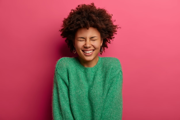 La niña de pelo rizado emocional feliz sonríe, expresa emociones positivas, cierra los ojos de la risa, usa un suéter verde, se siente optimista, aislada en la pared rosa, irradia felicidad.