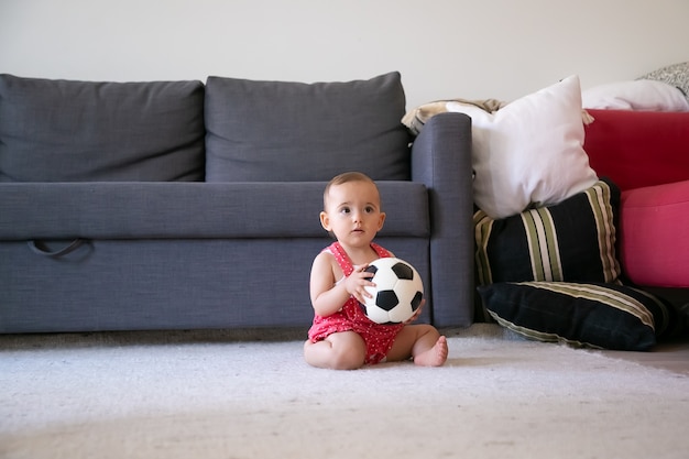 Niña pensativa sosteniendo un balón de fútbol, sentada sobre una alfombra descalza y mirando a otro lado. Niño encantador en pantalones cortos de peto rojo jugando en casa cerca del sofá. Concepto de vacaciones, fin de semana e infancia.