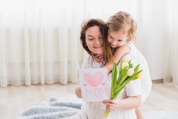 Niña pequeña abrazando a su madre desde atrás mientras sonríe madre sosteniendo una tarjeta de felicitación y flores en casa