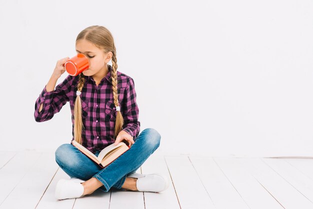 Niña pequeña adorable leyendo un libro y sosteniendo una taza