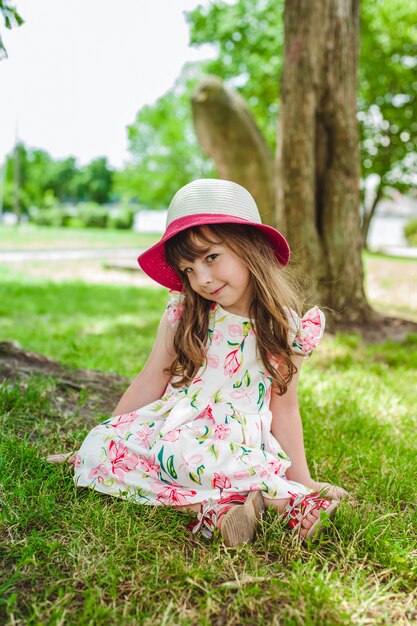 Niña pequeña adorable sentada en el suelo de un parque con un sombrero