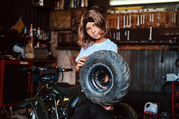 Foto gratuita una niña pequeña y agradable posa para el fotógrafo con una rueda grande del automóvil en el servicio de automóviles.