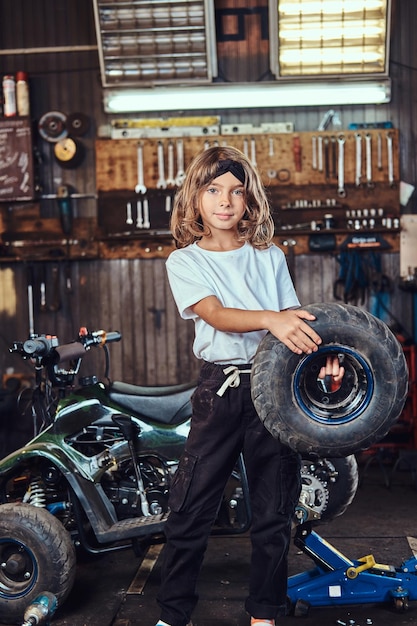 Una niña pequeña y agradable posa para el fotógrafo con una rueda grande del automóvil en el servicio de automóviles.