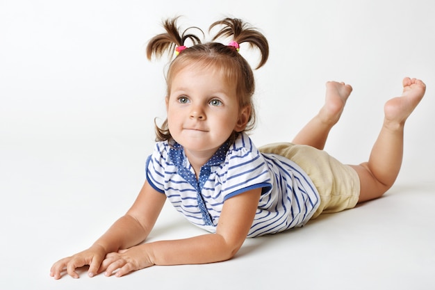 Niña pequeña con aspecto atractivo, expresión soñadora, tiene dos colas de caballo divertidas, levanta las piernas hacia arriba