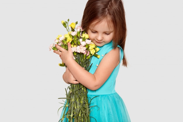 Niña pequeña, enfocada hacia abajo, vestida con un elegante vestido, lleva un ramo de flores de primavera, posa en blanco. Adorable niña recibe flores el 8 de marzo.