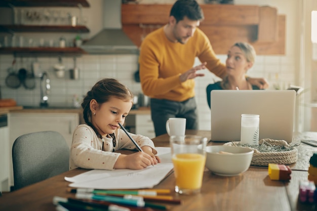 Niña pequeña escribiendo la tarea mientras los padres trabajan en segundo plano en casa