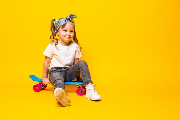 Niña pequeña con estilo en casual con patineta sobre pared amarilla.