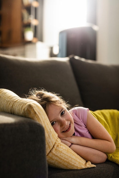 Niña pequeña feliz descansando en el sofá de la sala de estar y mirando a la cámara