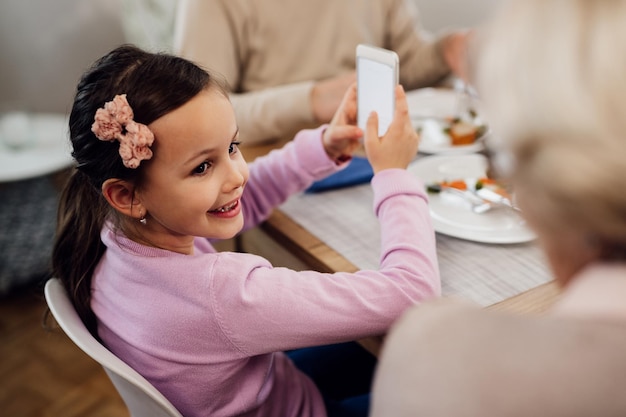 Niña pequeña feliz usando teléfono móvil y hablando con su abuela durante la hora del almuerzo en la mesa del comedor