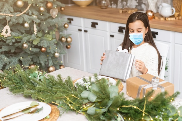 Niña pequeña con máscara protectora en el fondo del árbol de Navidad. Cara de niño triste con mascarilla quirúrgica. Navidad festiva y distanciamiento social durante el concepto de pandemia covid-19.