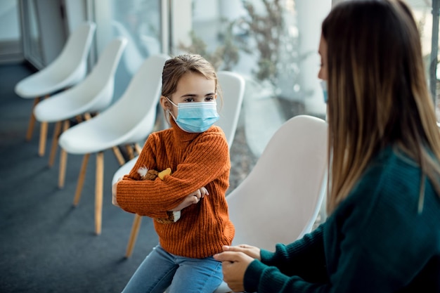 Niña pequeña con mascarilla haciendo pucheros mientras está con su madre en la sala de espera del hospital