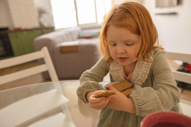 Niña pequeña de piel clara con cabello rojo en un bocadillo mientras se sienta a la mesa durante el día Concepto de nutrición