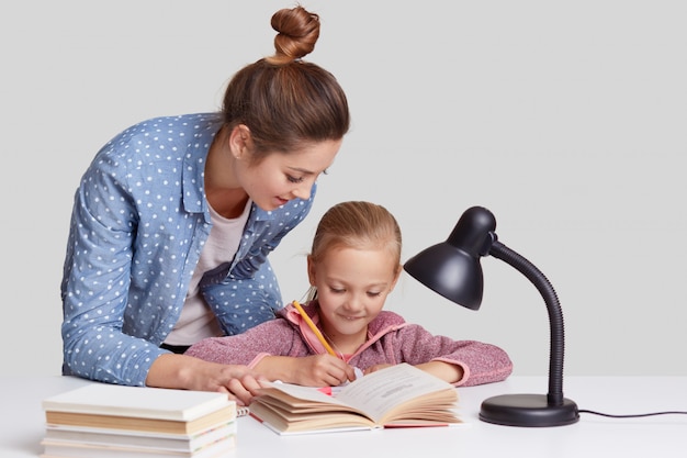 La niña pequeña reescribe la información en el cuaderno, tiene una expresión de satisfacción, su madre está cerca, trata de alentar a su hija para que estudie, ayude y explica el material, aislado en blanco