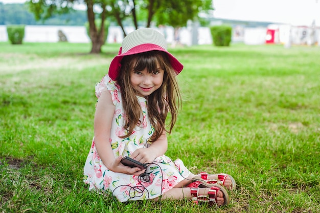 Niña pequeña sentada en el césped mirando un móvil con auriculares