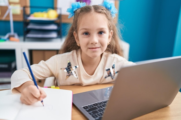 Foto gratuita niña pequeña sentada en la mesa haciendo la tarea con la computadora portátil mirando positiva y feliz de pie y sonriendo con una sonrisa confiada mostrando los dientes