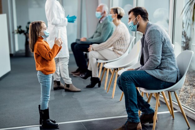 Una niña pequeña tomando una foto de su padre con un panel táctil en la sala de espera del hospital