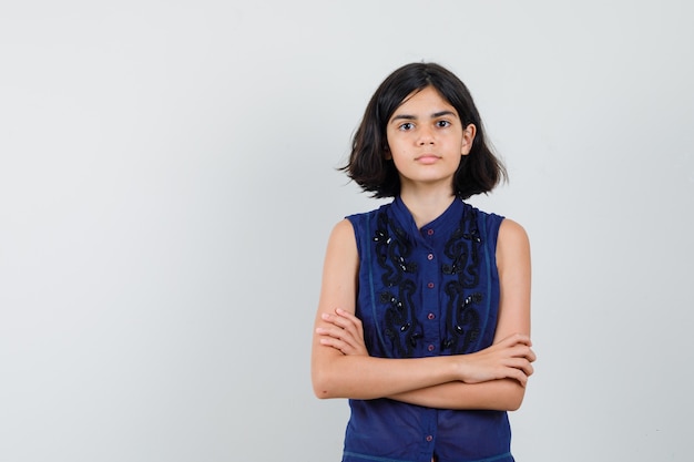 Niña de pie con los brazos cruzados en blusa azul y mirando serio