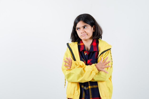 Niña de pie con los brazos cruzados en camisa a cuadros, chaqueta y mirando vacilante, vista frontal.