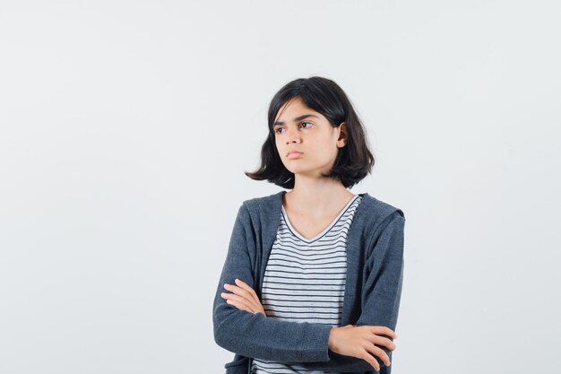 Niña de pie con los brazos cruzados en camiseta, chaqueta y mirando pensativo.