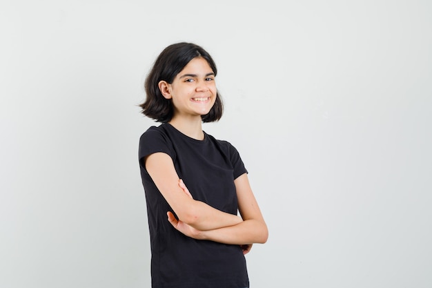 Niña de pie con los brazos cruzados en camiseta negra y mirando alegre, vista frontal.