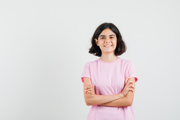 Niña de pie con los brazos cruzados en camiseta rosa y mirando alegre, vista frontal.