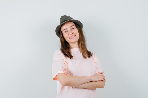 Niña de pie con los brazos cruzados en el sombrero de camiseta rosa y luciendo optimista