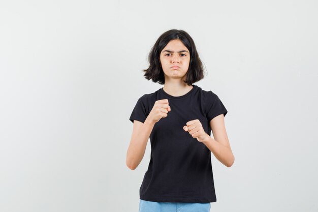 Niña de pie en pose de boxeador en camiseta negra, pantalones cortos y mirando enojado, vista frontal.