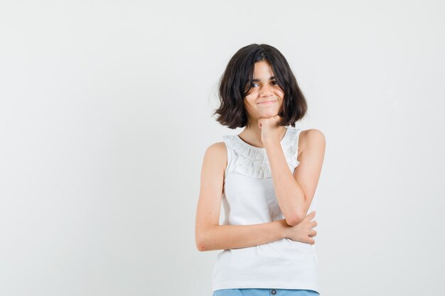 Niña de pie en pose de pensamiento en blusa blanca, pantalones cortos y bonita vista frontal.