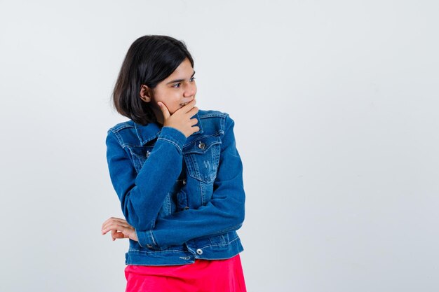 Niña de pie en pose de pensamiento en camiseta roja y chaqueta de jean y mirando pensativo