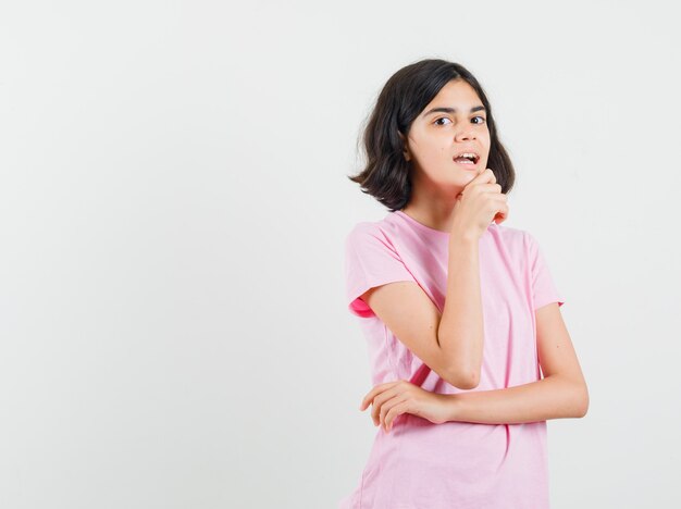 Niña de pie en pose de pensamiento en camiseta rosa y mirando curioso. vista frontal.