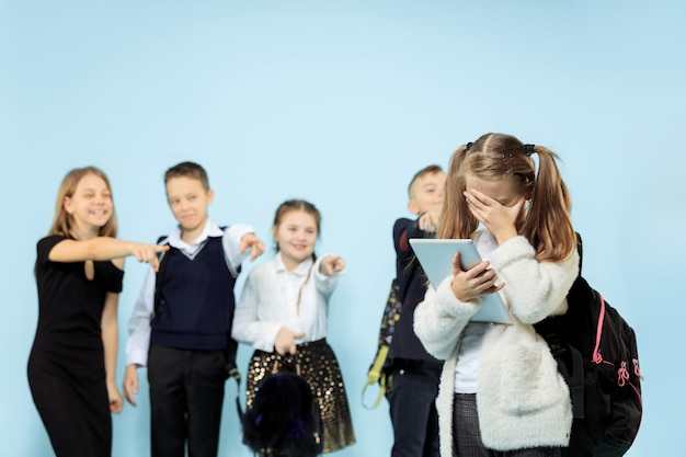 Foto gratuita niña de pie sola y sufriendo un acto de acoso mientras los niños se burlan. triste colegiala joven sentada en el estudio sobre fondo azul.