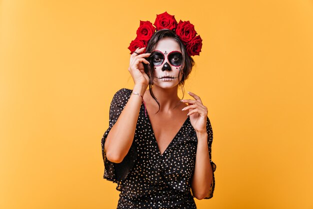Niña de piel oscura con corona de flores y máscara de calavera posa para la foto en memoria de Halloween. Retrato de modelo extraordinario en atuendo inusual