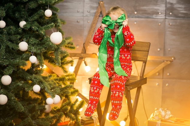 Niña en pijama junto al árbol de Navidad en una silla de madera