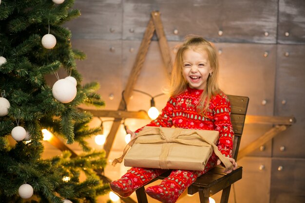 Niña en pijama junto al árbol de Navidad en una silla de madera
