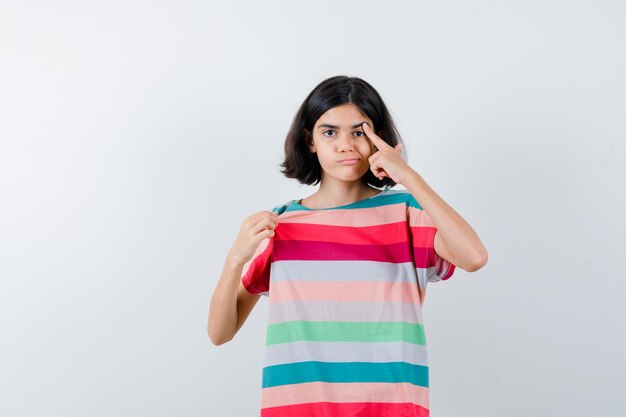 Niña poniendo el dedo índice en el ojo, curvándose los labios en camiseta, jeans y mirando serio, vista frontal.