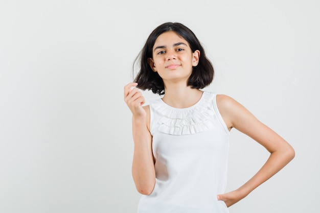 Niña posando mientras está de pie en una blusa blanca y parece optimista. vista frontal.