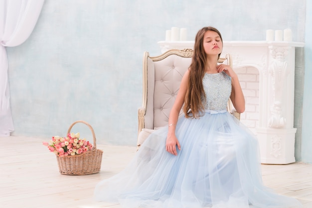 Niña posando mientras está sentado en el sillón cerca de la cesta de flores