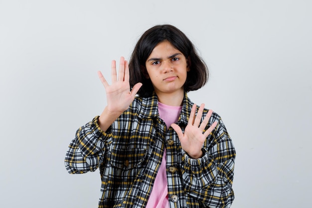 Niña preadolescente en camisa, chaqueta levantando las manos para defender y lucir insatisfecha, vista frontal.