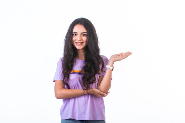 Niña presentando algo con la mano en la pared blanca y sonriendo.