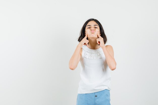 Niña presionando los dedos en las mejillas sopladas en blusa blanca, pantalones cortos y luciendo linda vista frontal.