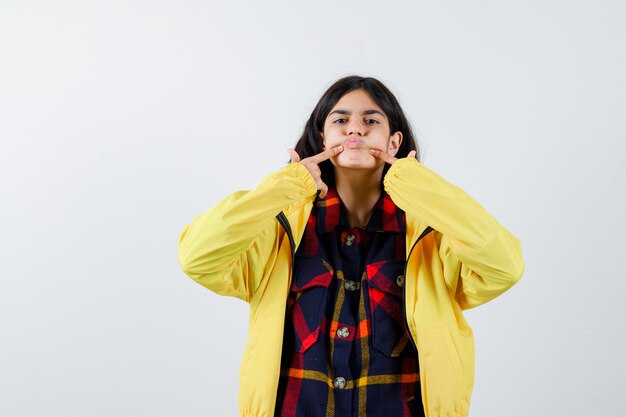 Niña presionando las mejillas con el dedo en camisa a cuadros, chaqueta y aspecto lindo