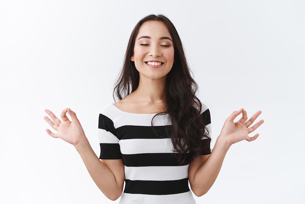 Foto gratuita la niña que controla sus pensamientos se mantiene positiva y relajada cada vez que cierra los ojos, respira profundamente y sonríe aliviada mientras medita, toma las manos de lado en el signo mudra zen, libera el estrés a través del yoga