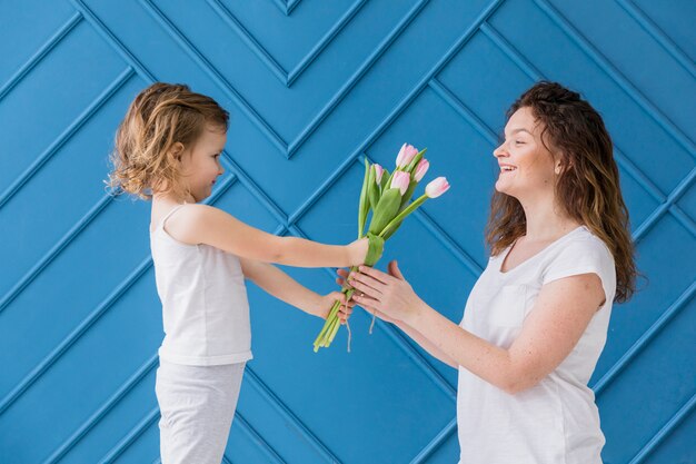 Niña que da flores rosadas de los tulipanes a su mamá en el día de madre delante del fondo azul