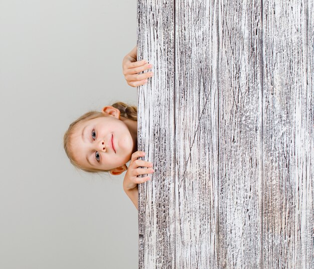 Niña que se esconde detrás del tablón de madera y que parece confiada.