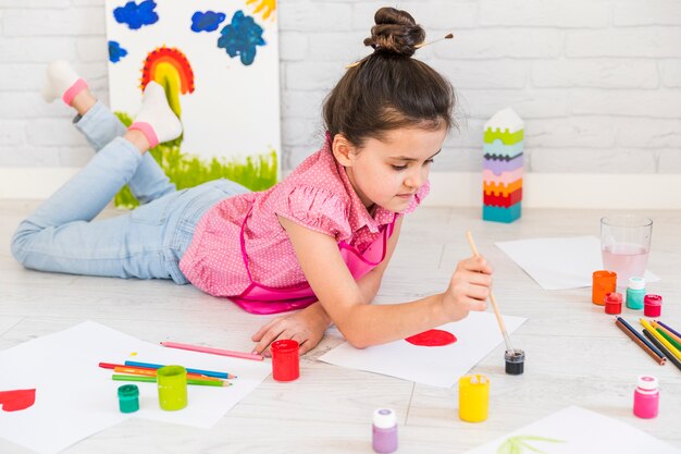 Niña que miente en la pintura del suelo con color de agua con el cepillo de pintura en el papel